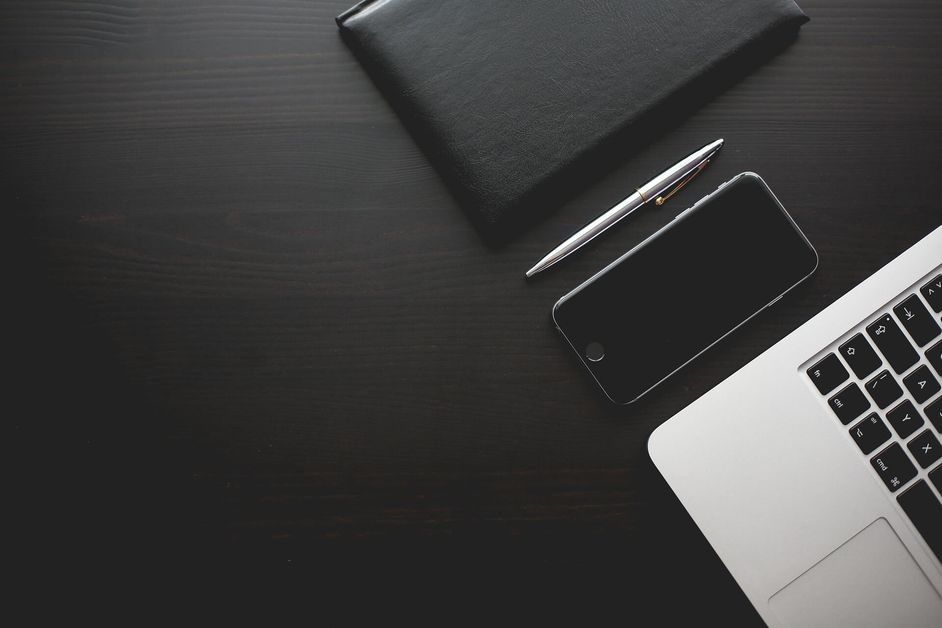 A dark wooden desk with a notebook, pen, phone, and laptop.